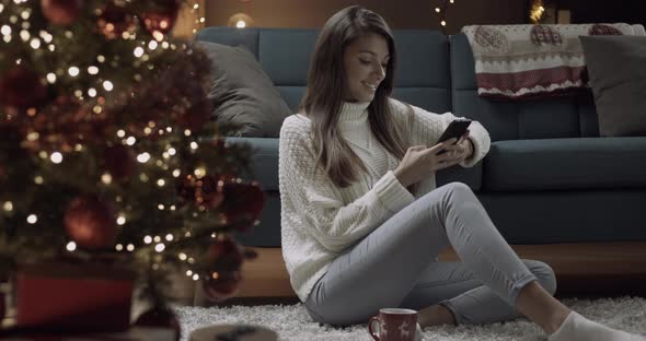 Young woman typing on a smartphone sitting near christmas tree and gifts.