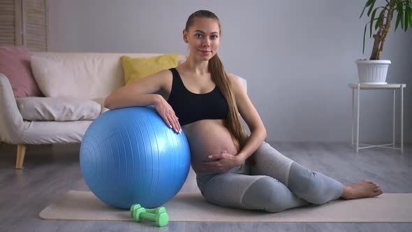 Pregnant Woman Practicing Yoga and Smiling While Sitting on Floor in Home Room Spbd alt