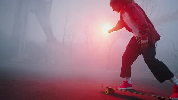 Trendy Teen Guy is Riding a Skateboard in the Evening Holding a Red Burning Torch in One Hand alt