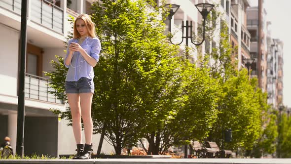 Young Beautiful Blonde Girl in Summer Day Walks at Urban City Park Street Listening Music with