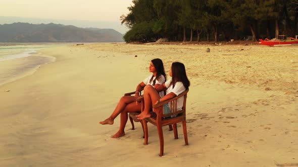 Two girls facing sea, sitting on chairs over white sand of exotic beach on a quiet sunset scenery of alt