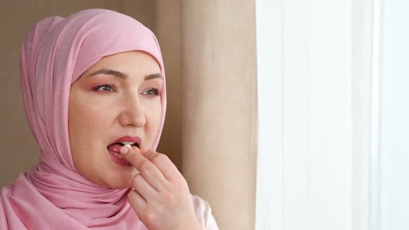 Young Woman in Hijab Drinks White Capsule with Water While Standing By the Window alt