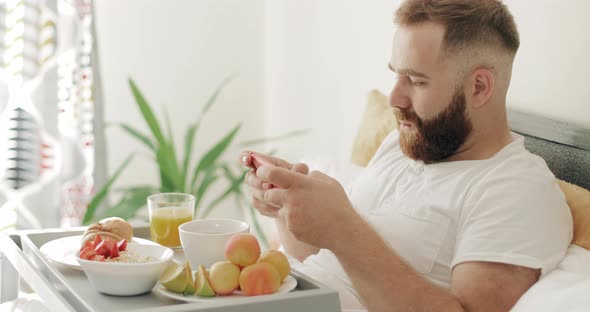 Side View of Young Guy Sitting on Bed and Playing Game on smartphone, Bearded Man in 30s Using  alt