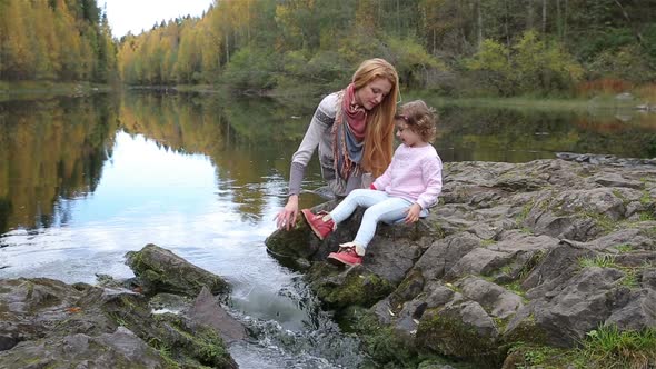 A Young Mother and Her Cute Daughter Sitting Near the Forest Stream alt