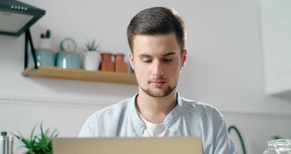 Young Bearded Man Works Home on Laptop in Kitchen, Drinking Coffee in Morning