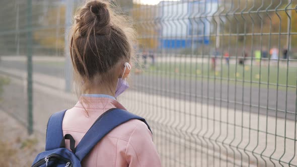 A Sad Girl in a Medical Mask Looks at Children Playing Sports on the Playground
