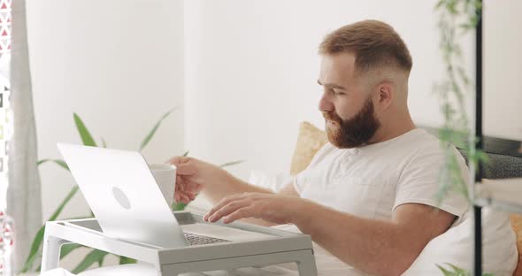 Side View of Good Looking Guy Typing and Smiling While Sitting on Bed in Morning, Bearded Male alt