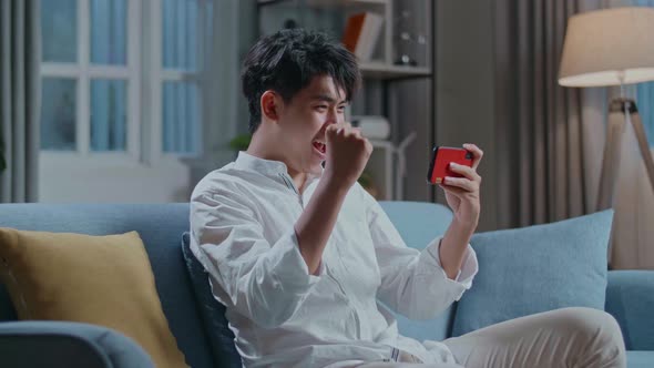Asian Man Celebrating Winning Game On Smartphone While Sitting On Sofa In The Living Room alt