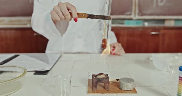 Solemn Young Female Chemistry Teacher in White Lab Coat and Glasses Heating Test Tube with Dark Red alt