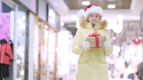 Little Girl in Santa Hat with Gifts Box in Christmas Walking in Shopping Mall Traditional Market alt
