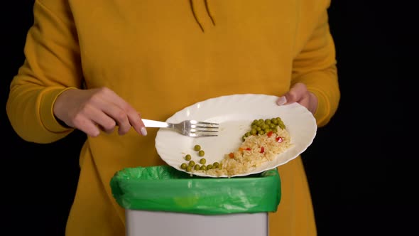Woman Scraping with a Plate a Chicken Leg, Rice, Green Peas Into Garbage Bin alt