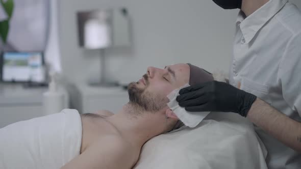 Side View of Relaxed Young Handsome Man Lying on Beauty Bed As Unrecognizable Beautician in Covid19 alt