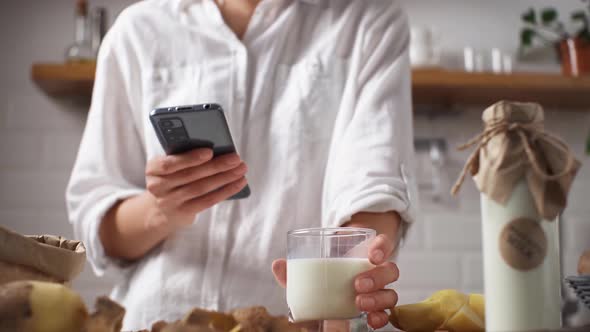 Girl With A Phone Is Standing In The Kitchen Drinking Vegetable Milk From Potatoes A Girl Is Looking alt