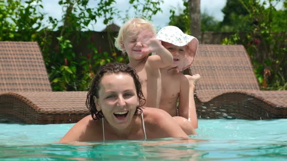 Cheerful Family of Young Mother and Toddler Twin Sisters Swimming in Pool Waving Hello to the Camera alt
