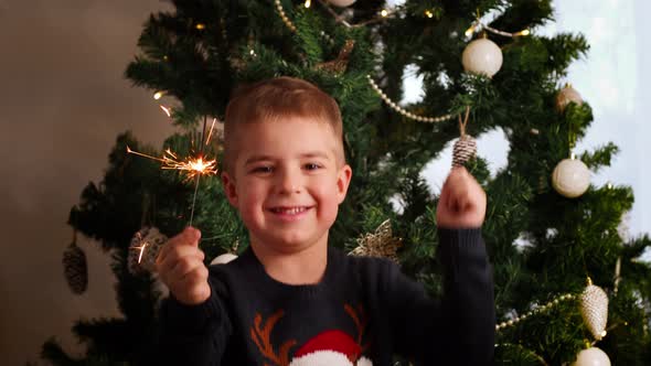 Little caucasian boy with Xmas sweater dancing by Christmas tree at home and holding sparkler