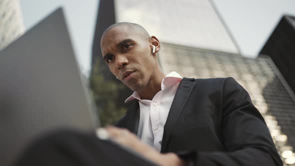 Displeased African businessman working on laptop and talking in headphones alt