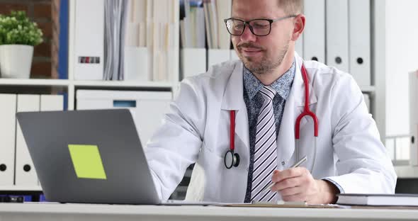 Young Doctor with Glasses Looking at Laptop Screen and Writing in Medical Documents  Movie alt
