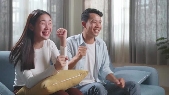 Excited Asian Couple Watching Tv Together Sitting On A Sofa In The Living Room At Home alt
