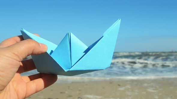 Person Holding Blue Paper Boat in Hand on Background of Sea Waves on Sunny Day alt