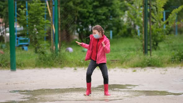 A little girl in a medical mask and rubber boots jumps through puddles on a spring day after rain