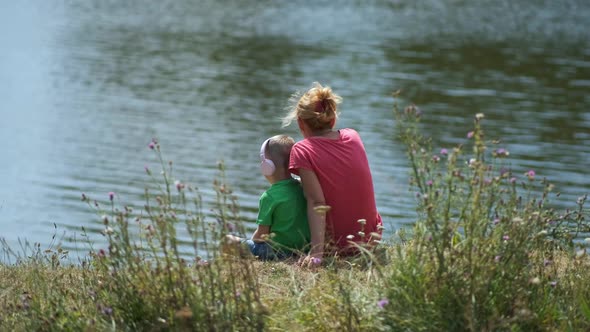 A mother and her young son are sitting on the shore of a lake. the boy listens to music