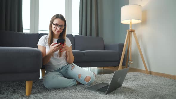 Casually Dressed Woman Sitting on Carpet with Laptop and Smartphone and Working in Cozy Room alt