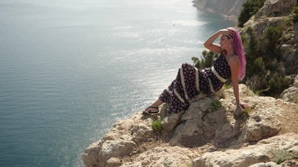 A Bright Girl in a Summer Dress Sits on Top of a Mountain Overlooking the Sea