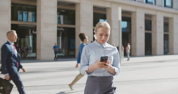 Caucasian Pretty Businesswoman Walking Outside Office Center Taping on Cellphone alt