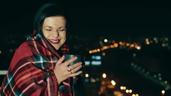 Woman on a Cold Night with a Cup of Tea on the Terrace of a Highrise Building alt