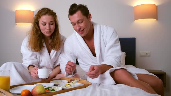 Man and Woman in Bathrobes Having a Breakfast Together in Bed in Hotel Room