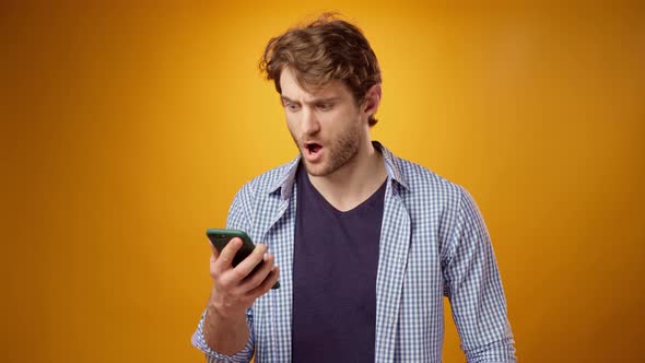 Young Man Looks at Cell Phone Screen with Astonishment and Disappointment Yellow Background alt