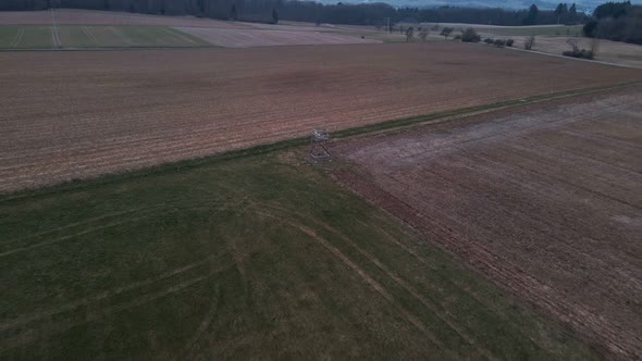 A hunter's tree stand in the middle of green and brown meadows in Germany during sunset. Fast aerial alt