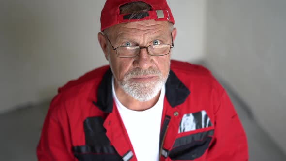 Portrait of an Elderly Engineer Worker in a Red Overalls and a Cap with a Gray Beard and Glasses alt