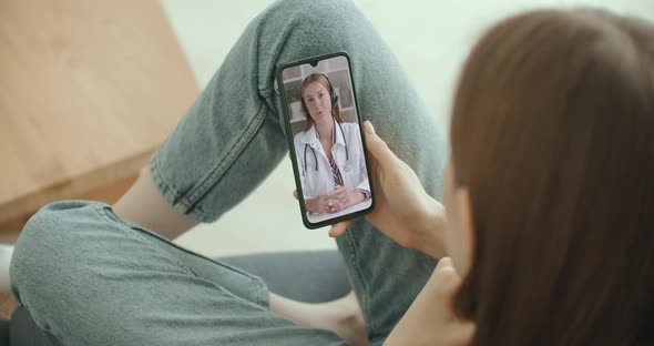 Female Medical Assistant Wears White Coat, Headset Video Calling Distant Patient on Smartphone alt