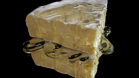 Drops of honey dripping from Brie cheese on a mirrored black background. alt