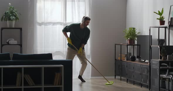 Joyful Man Is Dancing with Mop in Apartment Listening To Music By Headphones and Singing Song alt