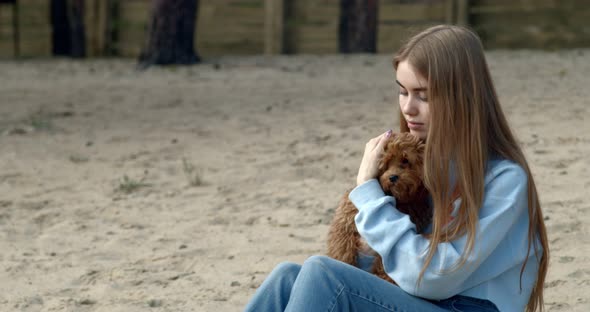 A Young Girl is Sitting on the Shore of a Forest Lake with a Little Puppy and Hugging Him