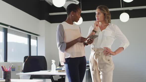 Two diverse female colleagues looking at tablet and discussing in office alt