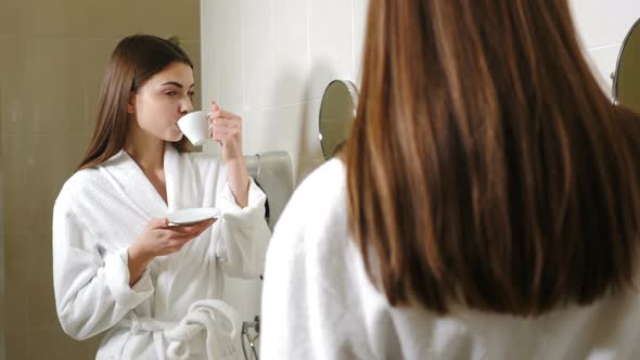 Young Attractive Woman in White Bathrobe Drinking Coffee in Front of Bathroom Mirror alt