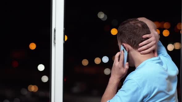 Young Man Talking on the Phone Standing at the Open Window at Night Rear View alt