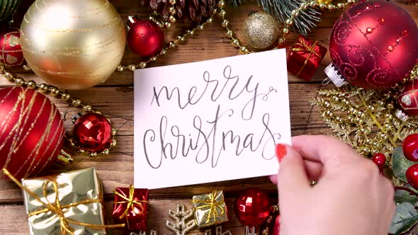 Woman's hand putting a card with the text MERRY CHRISTMAS on a wooden table alt