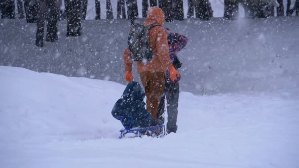 Family of Dad, Mom, Little Son and Daughter Riding on a Sled in a Pine Forest in Snowfall alt