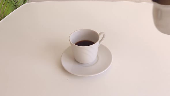A Woman Pours Milk From a Metal Ladle Into a White Cup of Coffee Standing on a White Table