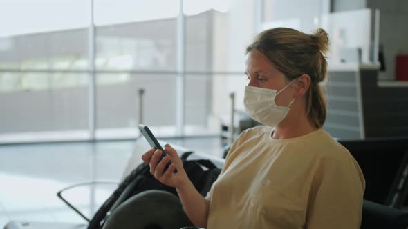Woman Wearing Medical Mask Protection and Using Smartphone While Sitting in Airport Terminal and alt