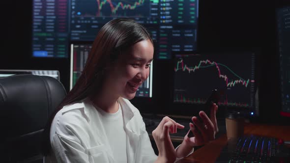 Asian Female  Stock Market Broker Using Mobile Phone With Analyzing Graphs On Multiple Computer alt