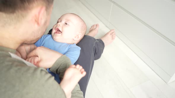 Father Plays with a Smiling Baby Son Lying on Knees at Home Together