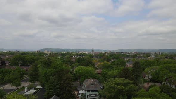 Flyover of small city neighborhood with tree lined streets, clouds, mountains in distance.