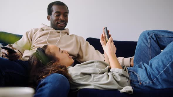 Relaxed Young Couple At Home Sitting On Sofa Watching TV And Checking Social Media On Mobile Phone