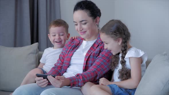Mother Playing with Children at Home Relaxing Use a Smartphone Cuddling Sit on Sofa Daughter and Son alt