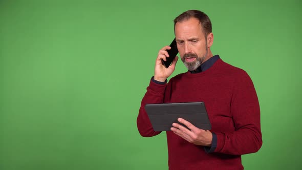 A Middleaged Handsome Caucasian Man Talks on a Smartphone and Holds a Tablet  Green Screen